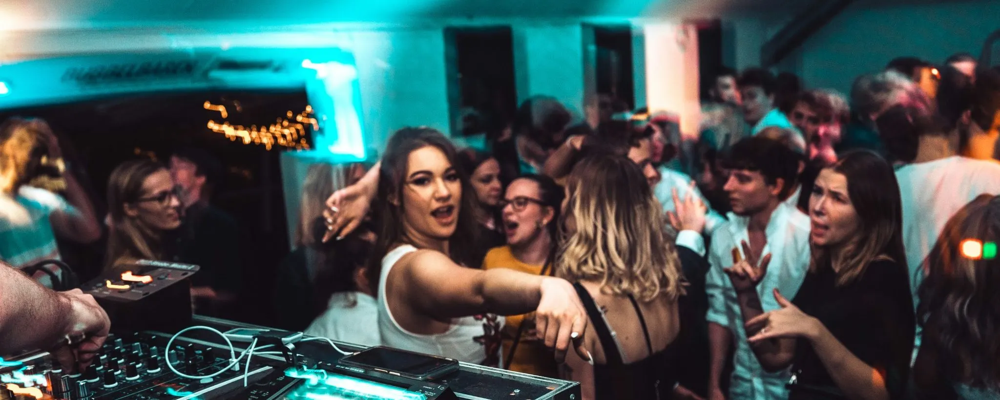 A small room packed tight beside the booth, the decks lit in the foreground and people dancing close behind them
