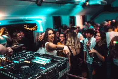 A small room packed tight beside the booth, the decks lit in the foreground and people dancing close behind them